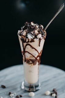 A tall glass is filled with a creamy beverage topped with whipped cream, chocolate drizzle, and crumbled cookie pieces. A spoon is placed in the glass, with some chocolate pieces scattered on the marble surface below.