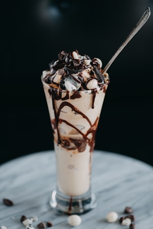 A tall glass is filled with a creamy beverage topped with whipped cream, chocolate drizzle, and crumbled cookie pieces. A spoon is placed in the glass, with some chocolate pieces scattered on the marble surface below.