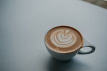 A white ceramic cup filled with a latte featuring intricate latte art on a smooth, light-colored surface. The design resembles a heart and leaf pattern. The background is a muted gray, providing a minimalist and calm setting.