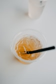 A close-up of a plastic cup filled with an amber-colored iced drink and a black straw, set on a minimalistic white surface. Another translucent cup, possibly filled with water, is partially visible in the background.