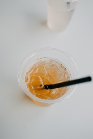A close-up of a plastic cup filled with an amber-colored iced drink and a black straw, set on a minimalistic white surface. Another translucent cup, possibly filled with water, is partially visible in the background.