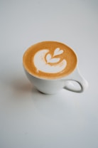 A cup of coffee with intricate latte art on top, featuring a creamy white design set against a rich brown background. The cup is white and sits on a smooth, light-colored surface, casting a soft shadow.