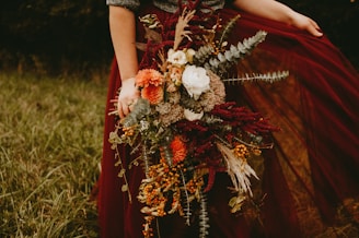 A person holds a large, rustic bouquet featuring a variety of autumnal flowers and foliage. The arrangement includes white roses, orange chrysanthemums, branches of berries, and dried leaves, all complemented by a deep burgundy skirt or fabric.