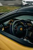 The interior of a car is shown with a focus on the steering wheel featuring a recognizable emblem. The dashboard includes modern digital displays and control buttons. The view is partially obscured by the car's exterior, which is in a bright yellow color. In the background, part of another vehicle is visible in a parking lot setting.