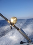 A fishing rod with a gold reel is mounted on a holder as a boat speeds over the ocean, causing splashes of water and creating a sense of motion. The sky above is clear and blue, and a rainbow is faintly visible in the spray.