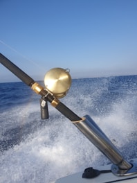A fishing rod with a gold reel is mounted on a holder as a boat speeds over the ocean, causing splashes of water and creating a sense of motion. The sky above is clear and blue, and a rainbow is faintly visible in the spray.