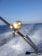 A fishing rod with a gold reel is mounted on a holder as a boat speeds over the ocean, causing splashes of water and creating a sense of motion. The sky above is clear and blue, and a rainbow is faintly visible in the spray.