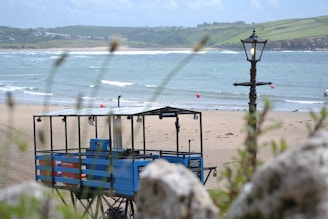 A coastal scene featuring a sandy beach with gentle waves in the background. In the foreground, a rustically designed lamp post stands near a blue lifeguard station, which is elevated on stilts. The scenery includes rolling green hills in the distance, contributing to a serene seaside atmosphere.