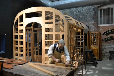 A workshop scene featuring a mannequin of a carpenter working on the wooden frame of a structure. The setting includes various wooden components, tools, and ladders around the workspace. The environment has a rustic and industrial feel with exposed brick walls and a large metal duct overhead.