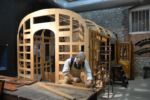 A workshop scene featuring a mannequin of a carpenter working on the wooden frame of a structure. The setting includes various wooden components, tools, and ladders around the workspace. The environment has a rustic and industrial feel with exposed brick walls and a large metal duct overhead.