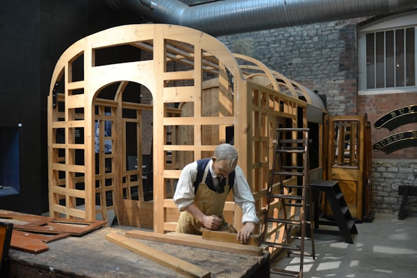 A workshop scene featuring a mannequin of a carpenter working on the wooden frame of a structure. The setting includes various wooden components, tools, and ladders around the workspace. The environment has a rustic and industrial feel with exposed brick walls and a large metal duct overhead.