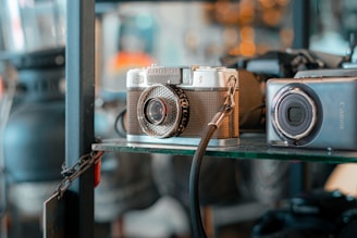 Two vintage cameras are displayed on a glass shelf. One camera has a retro design with a textured brown body and a silver lens, while the other is more compact with a sleek gray finish. Both cameras are tethered by cables, and blurred warm lights create a cozy atmosphere in the background.