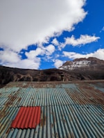 A corrugated metal roof dominates the foreground, with weathered and rusted patches and a single red sheet standing out among the blue-green panels. In the background, a rocky mountain partially covered in snow rises under a sky filled with fluffy white clouds.