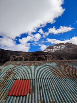 A corrugated metal roof dominates the foreground, with weathered and rusted patches and a single red sheet standing out among the blue-green panels. In the background, a rocky mountain partially covered in snow rises under a sky filled with fluffy white clouds.