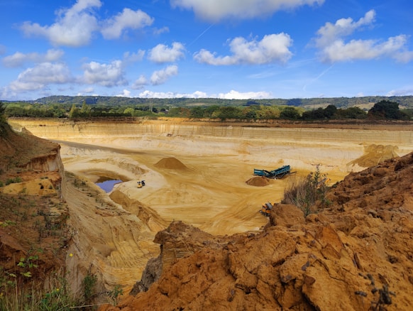 A large open quarry with sandy and rocky terrain, surrounded by green trees and hills in the distance. A blue industrial machine and a small construction vehicle are visible on the quarry floor. The sky is bright with scattered clouds.