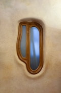 A uniquely shaped wooden-framed window with blue-tinted glass set in a textured wall. The window has an organic, asymmetrical form, reminiscent of natural shapes.