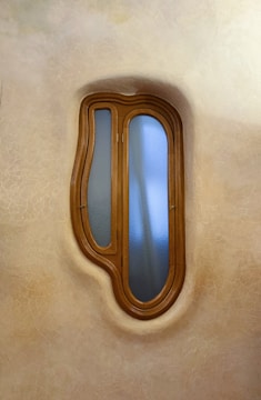 A uniquely shaped wooden-framed window with blue-tinted glass set in a textured wall. The window has an organic, asymmetrical form, reminiscent of natural shapes.