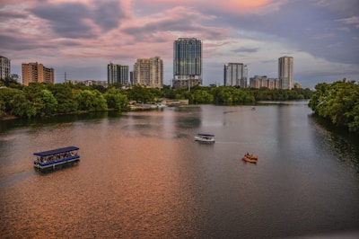A river with several small boats, including a large blue tour boat, glides through the water with dense trees lining the banks. Modern high-rise buildings dominate the skyline under a dramatic, colorful sky with soft hues of pink and orange during sunset.