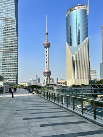 A modern urban scene with tall skyscrapers and the iconic Oriental Pearl Tower in the background. The scene is vibrant and busy, with pedestrians walking on a wide path beside a series of contemporary buildings.