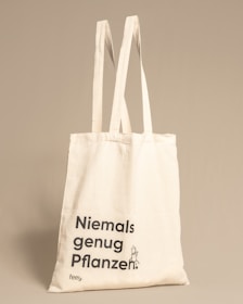 A beige tote bag with long handles featuring the text 'Niemals genug Pflanzen' printed in black, accompanied by a small illustration of a plant character.
