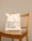 A beige reusable tote bag with black text, placed on a wooden chair with vertical slats on the backrest. The background is a plain light tan color.