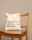 A beige reusable tote bag with black text, placed on a wooden chair with vertical slats on the backrest. The background is a plain light tan color.