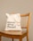 A beige reusable tote bag with black text, placed on a wooden chair with vertical slats on the backrest. The background is a plain light tan color.