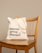 A beige reusable tote bag with black text, placed on a wooden chair with vertical slats on the backrest. The background is a plain light tan color.