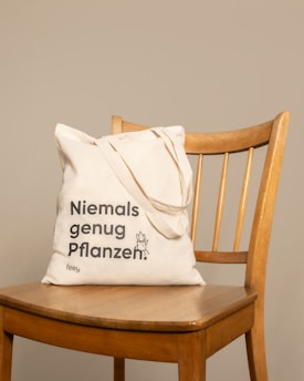 A beige reusable tote bag with black text, placed on a wooden chair with vertical slats on the backrest. The background is a plain light tan color.