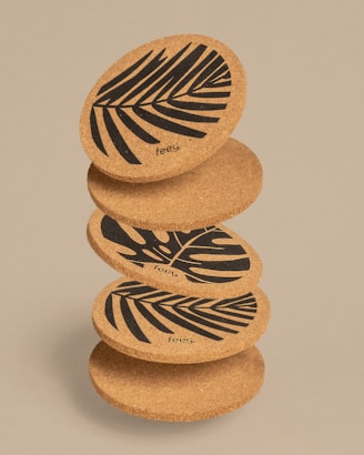 A stack of round cork coasters, each decorated with a bold black leaf pattern. The design features stylized tropical leaves, and the coasters are positioned in a slightly tilted manner, displaying the artwork prominently.