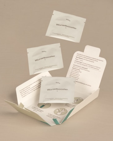 The image features a neutral-toned background with several sachets labeled 'Wurzelbooster' floating above and inside an open package. The package includes printed instructions and illustrations, suggesting a product meant for enhancing root growth. The sachets and packaging have a clean, minimalist design.