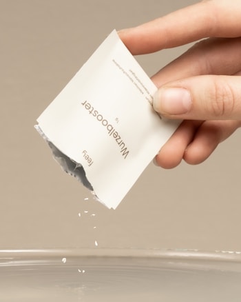 A hand holds a packet labeled 'WaterBooster' over a bowl. The packet is being opened, allowing its contents to fall into the water below.