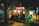 A small street food stall illuminated by warm lights, surrounded by urban cityscape at night. The stall has vibrant red curtains with Japanese lettering, offering a cozy and inviting atmosphere. Various items are stacked around, hinting at a busy, functional space.