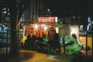 A small street food stall illuminated by warm lights, surrounded by urban cityscape at night. The stall has vibrant red curtains with Japanese lettering, offering a cozy and inviting atmosphere. Various items are stacked around, hinting at a busy, functional space.