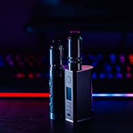 Two metallic vape devices are positioned in the foreground with a sleek design and buttons on their bodies. The background is a blur of colorful lights, featuring tones of blue and red, suggesting a digital or gaming environment.