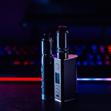 Two metallic vape devices are positioned in the foreground with a sleek design and buttons on their bodies. The background is a blur of colorful lights, featuring tones of blue and red, suggesting a digital or gaming environment.