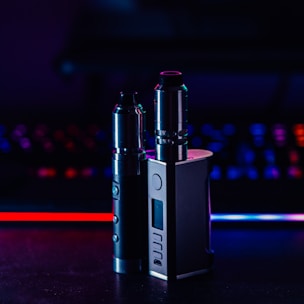 Two metallic vape devices are positioned in the foreground with a sleek design and buttons on their bodies. The background is a blur of colorful lights, featuring tones of blue and red, suggesting a digital or gaming environment.