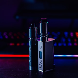 Two metallic vape devices are positioned in the foreground with a sleek design and buttons on their bodies. The background is a blur of colorful lights, featuring tones of blue and red, suggesting a digital or gaming environment.