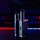 Two metallic vape devices are positioned in the foreground with a sleek design and buttons on their bodies. The background is a blur of colorful lights, featuring tones of blue and red, suggesting a digital or gaming environment.