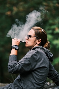 A person wearing a gray hoodie and glasses is sitting outdoors, exhaling a large cloud of vapor from a vaping device. The individual appears relaxed, with a watch on their wrist and hair tied back. The background is blurred with greenery suggesting a natural setting.