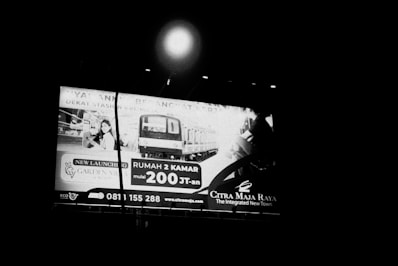 A large billboard is displayed in a nighttime setting, featuring advertisements for a real estate project. The sign includes an image of a residential area and a modern train. Contact information and a web address are also visible on the billboard.