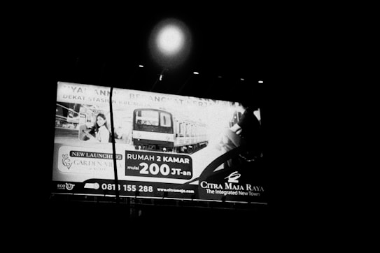 A large billboard is displayed in a nighttime setting, featuring advertisements for a real estate project. The sign includes an image of a residential area and a modern train. Contact information and a web address are also visible on the billboard.