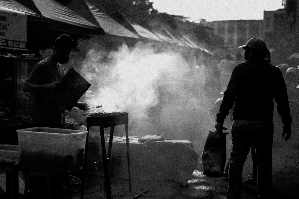a man standing next to another man cooking on a grill