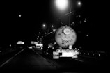 A large tanker truck is driving on a highway at night. The rear of the tanker is visible, with the number 20000 KG prominently displayed. Streetlights illuminate the scene, creating reflections on the wet road surface. Multiple vehicles can be seen in the background, and highway signs are visible along the road.