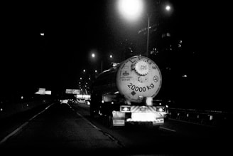 A large tanker truck is driving on a highway at night. The rear of the tanker is visible, with the number 20000 KG prominently displayed. Streetlights illuminate the scene, creating reflections on the wet road surface. Multiple vehicles can be seen in the background, and highway signs are visible along the road.