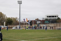 A soccer match is taking place on a grass field with players in blue and dark-colored uniforms. The scene includes a soccer goal with players lined in front of it, possibly preparing for a corner kick or similar play. A tall stadium light towers over the field, and there are various buildings and advertisements in the background. Flags are waving near the field, and a few vehicles are parked along the sidelines.