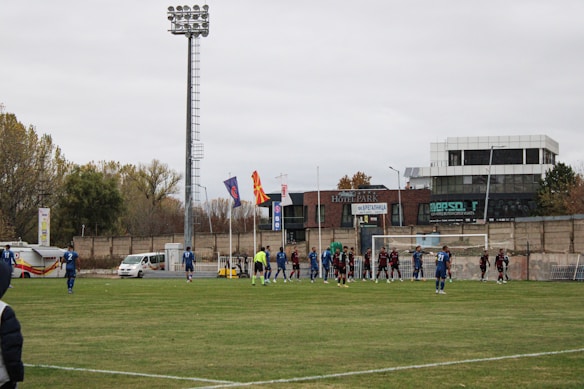 A soccer match is taking place on a grass field with players in blue and dark-colored uniforms. The scene includes a soccer goal with players lined in front of it, possibly preparing for a corner kick or similar play. A tall stadium light towers over the field, and there are various buildings and advertisements in the background. Flags are waving near the field, and a few vehicles are parked along the sidelines.