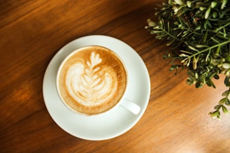 A steaming cup of latte art coffee beside a small vase with tropical leaves on a minimalist cafe table.