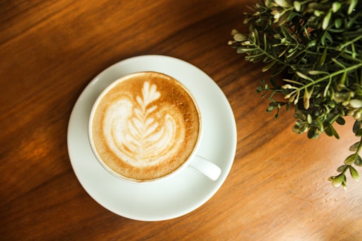 A steaming cup of latte art coffee beside a small vase with tropical leaves on a minimalist cafe table.