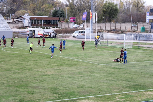 A football match is taking place on a grassy field with players wearing blue and black jerseys. One player lies on the ground while others are scattered around the field. There is an ambulance parked nearby, and flags are flying high. A referee in a bright uniform is present, and the background shows a few buildings and trees.